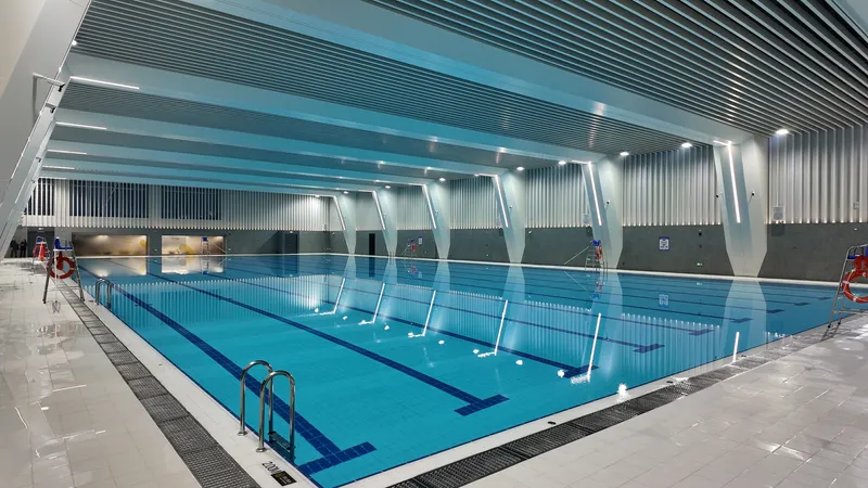 Indoor swimming pool with ribbed ceiling panels and angled white columns reflected in the water
