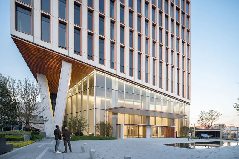 Glazed lobby entrance beneath a cantilevered timber soffit with two figures in the plaza at dusk