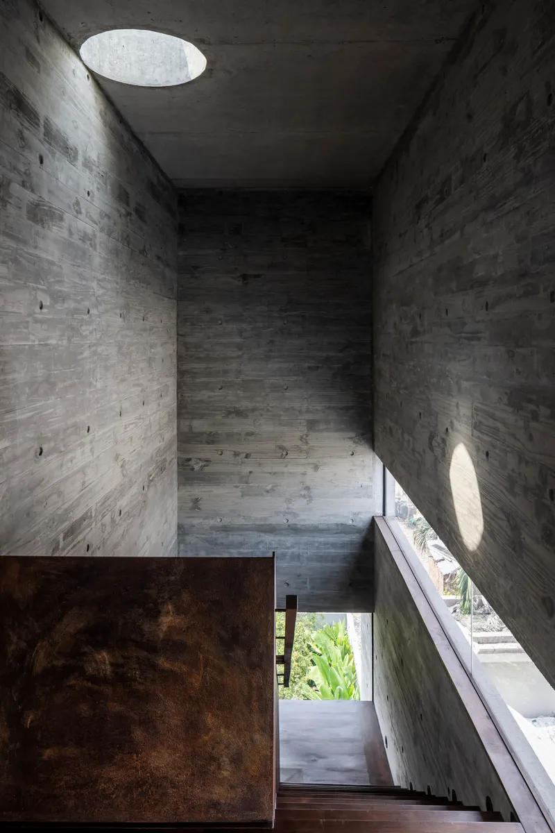 Board-formed concrete stairwell with circular ceiling oculus and angled shadows from afternoon sun