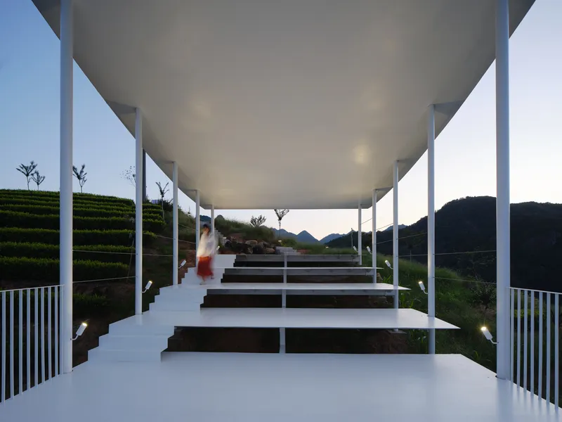 View through white steel columns supporting canopy above timber bench platforms at dusk with terraced landscape