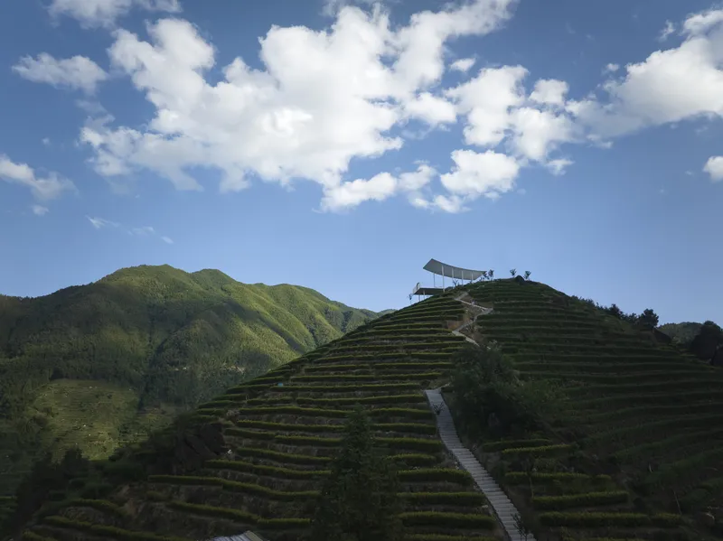 White canopy pavilion perched atop terraced hillside under blue sky with cumulus clouds and mountain backdrop