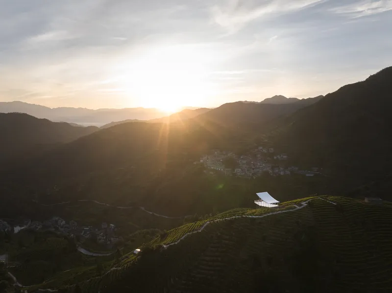 White pavilion on hillside plantation at sunset with distant mountain village in shadow