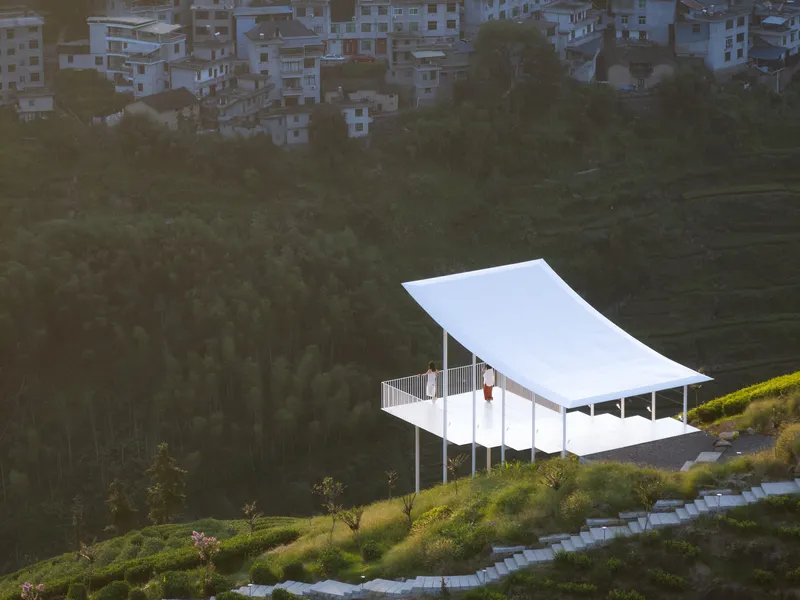 Elevated pavilion with curved white roof and slatted railing set among forested hillside with distant buildings