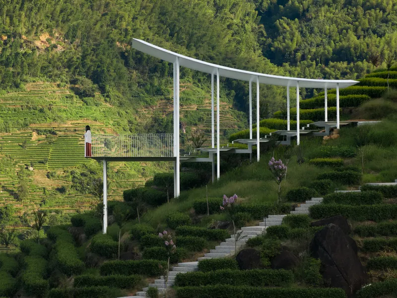 White steel canopy on slender columns spanning hillside terrace with visitor and flowering plants below