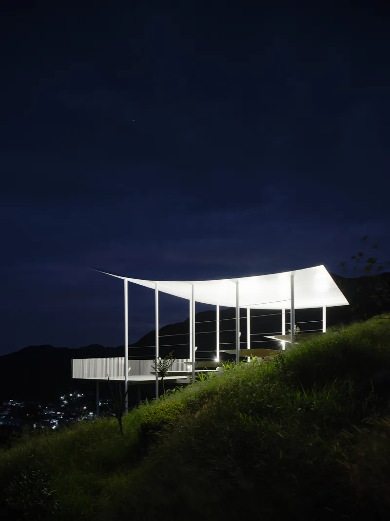 Side view of the illuminated pavilion with curved roof on a grassy slope at night