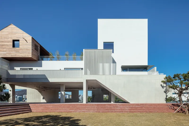 Elevated concrete structure with stacked volumes in timber, white corrugated metal, and stucco above a red brick platform and lawn