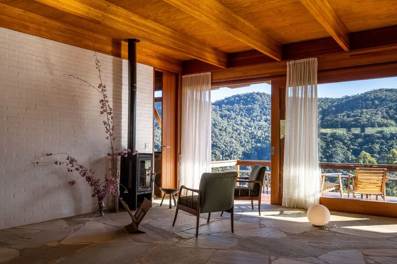 Living room with white brick fireplace, timber ceiling beams, and floor-to-ceiling windows overlooking forested valley
