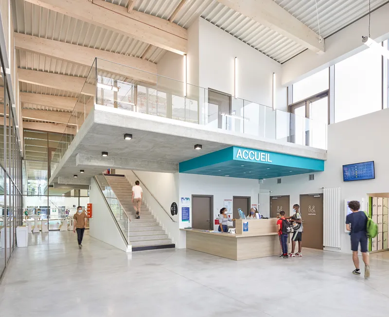 Double-height reception lobby with timber-slatted ceiling, mezzanine walkway, and visitors at the wooden desk below