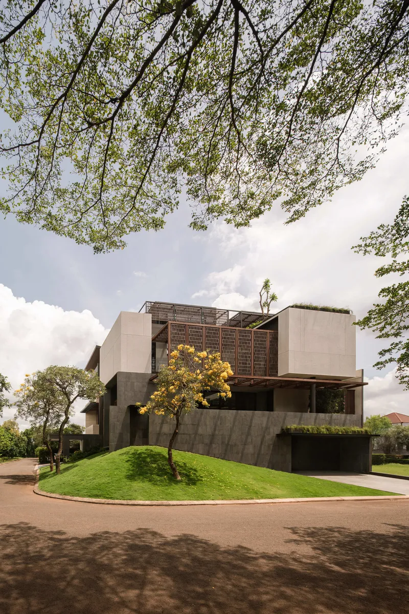 Street view of stacked volumes with lattice screens and yellow flowering tree on sloped lawn