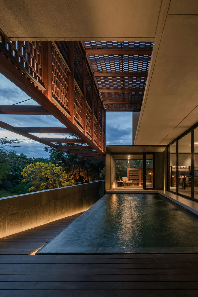 Timber deck pool at dusk with illuminated lattice canopy and glass-walled interior beyond