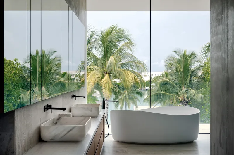 Bathroom interior with freestanding tub and dual stone basins opening to palm trees through floor-to-ceiling glass