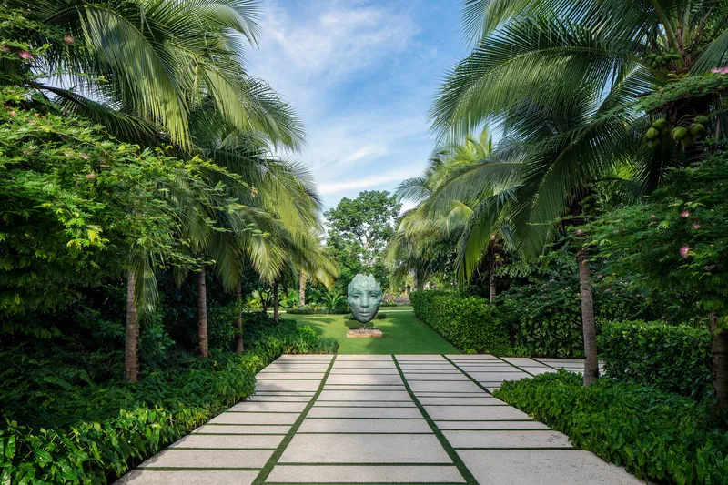 Paved garden path with grass joints leading to a sculptural head beneath palm canopy and blue sky