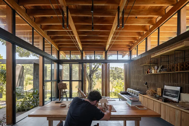 Open living space with timber ceiling beams, floor-to-ceiling glazing, and a person working at a table