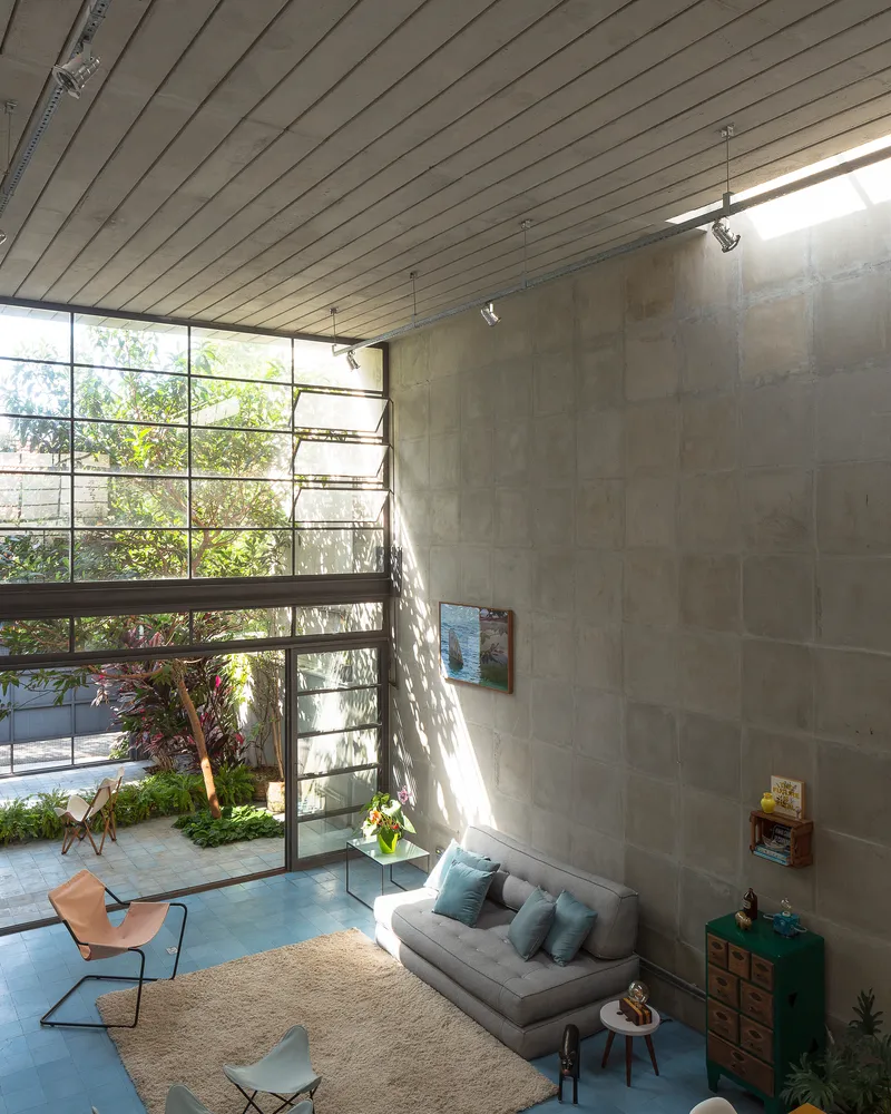 View from the upper mezzanine down into the living room with sunlight casting foliage shadows on concrete walls