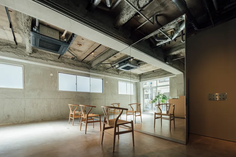 Glass-enclosed room with exposed concrete walls and ceiling ducts reflecting wooden chairs on polished floor