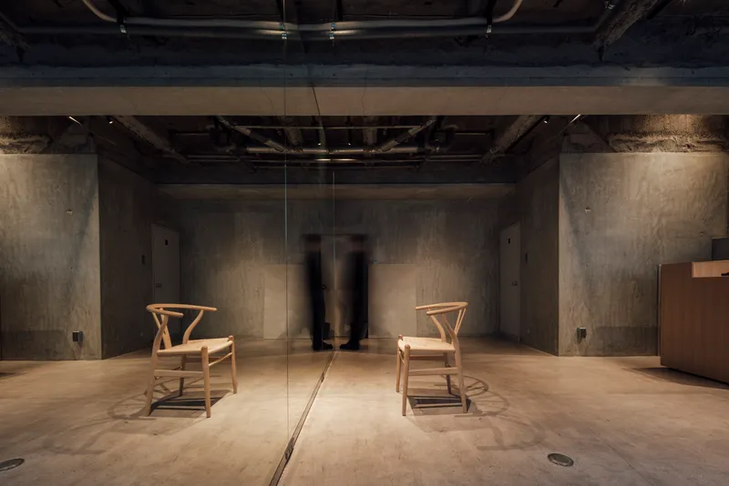 Glass partition reflecting two figures standing between wooden chairs in dimly lit concrete space