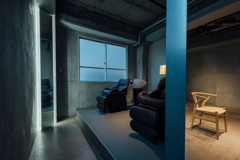Room with raw concrete walls, translucent glass windows, and reclined salon chairs under exposed services
