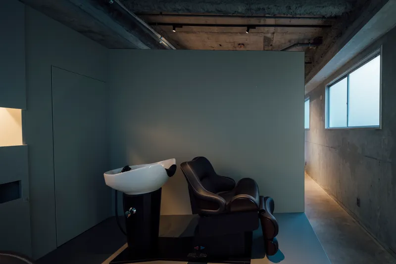 Salon space with wall-mounted washbasin, reclining chair, and concrete surfaces lit by natural light