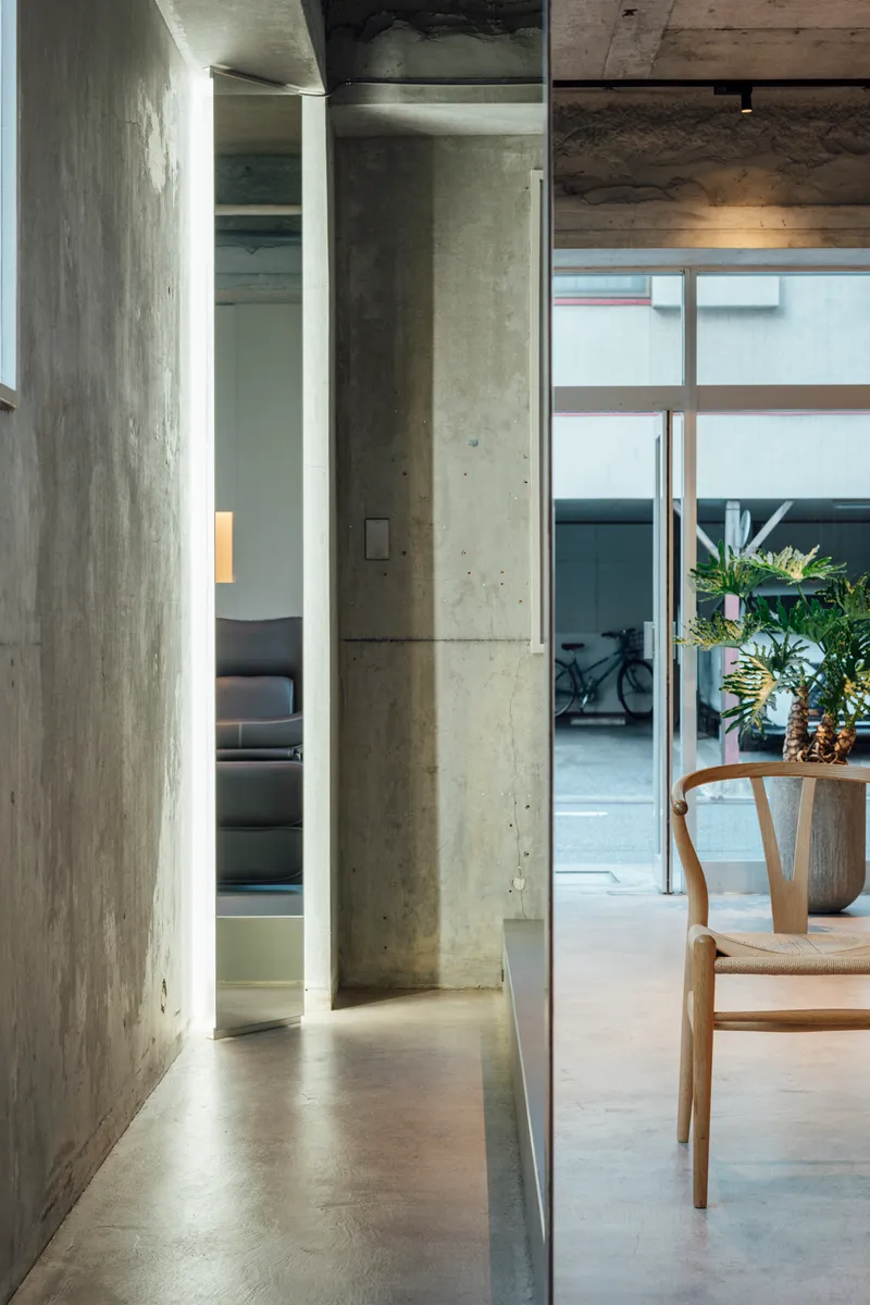 Corridor with exposed concrete walls and ceiling leading past a wooden chair to a glazed courtyard