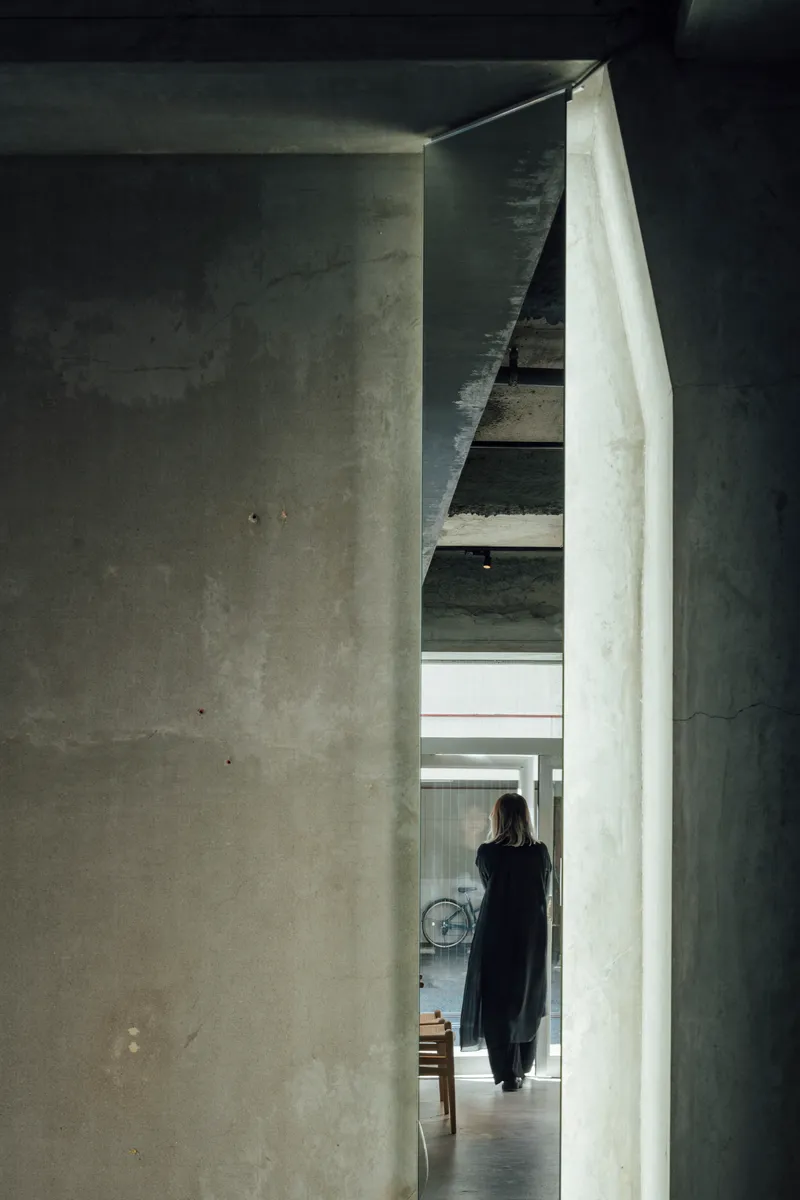 Narrow concrete passageway with a figure walking toward a staircase under indirect natural light