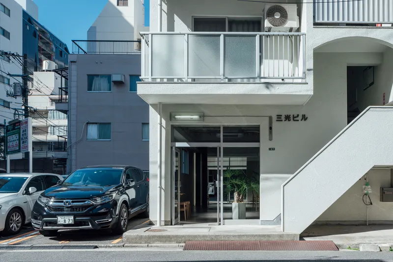 Street facade with glass entry doors and metal balcony railing above parked cars in daylight