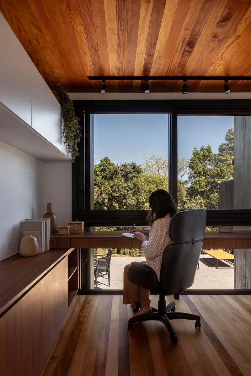 Home office with timber desk facing floor-to-ceiling windows overlooking green treetops in daylight