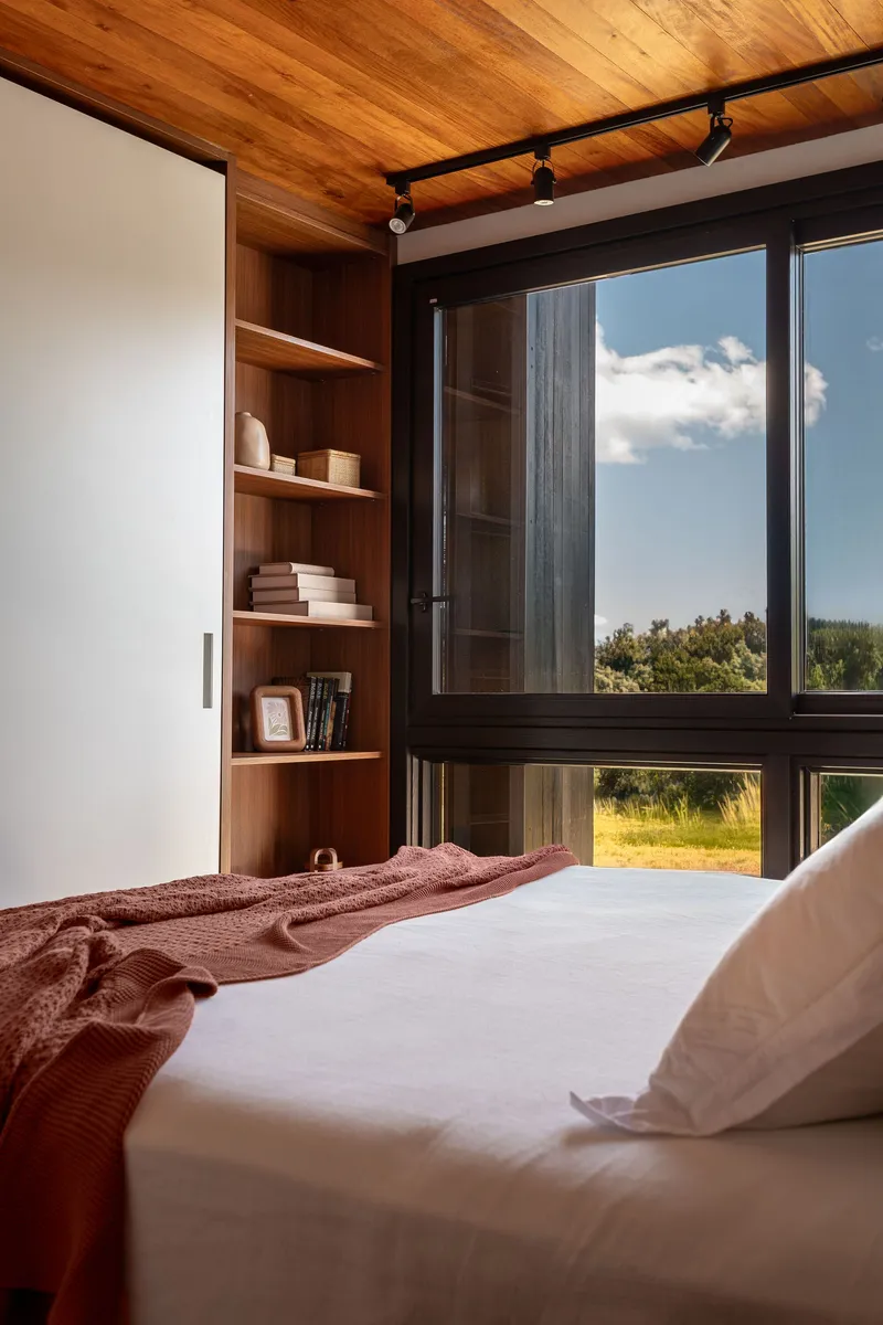Bedroom with built-in timber shelving beside black-framed window opening to landscape and sky