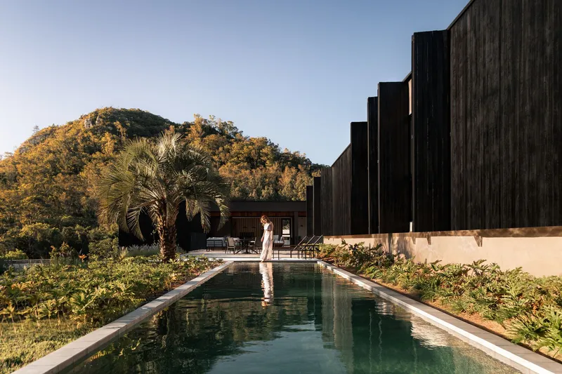 Long lap pool beside black timber volumes with a person walking along the deck