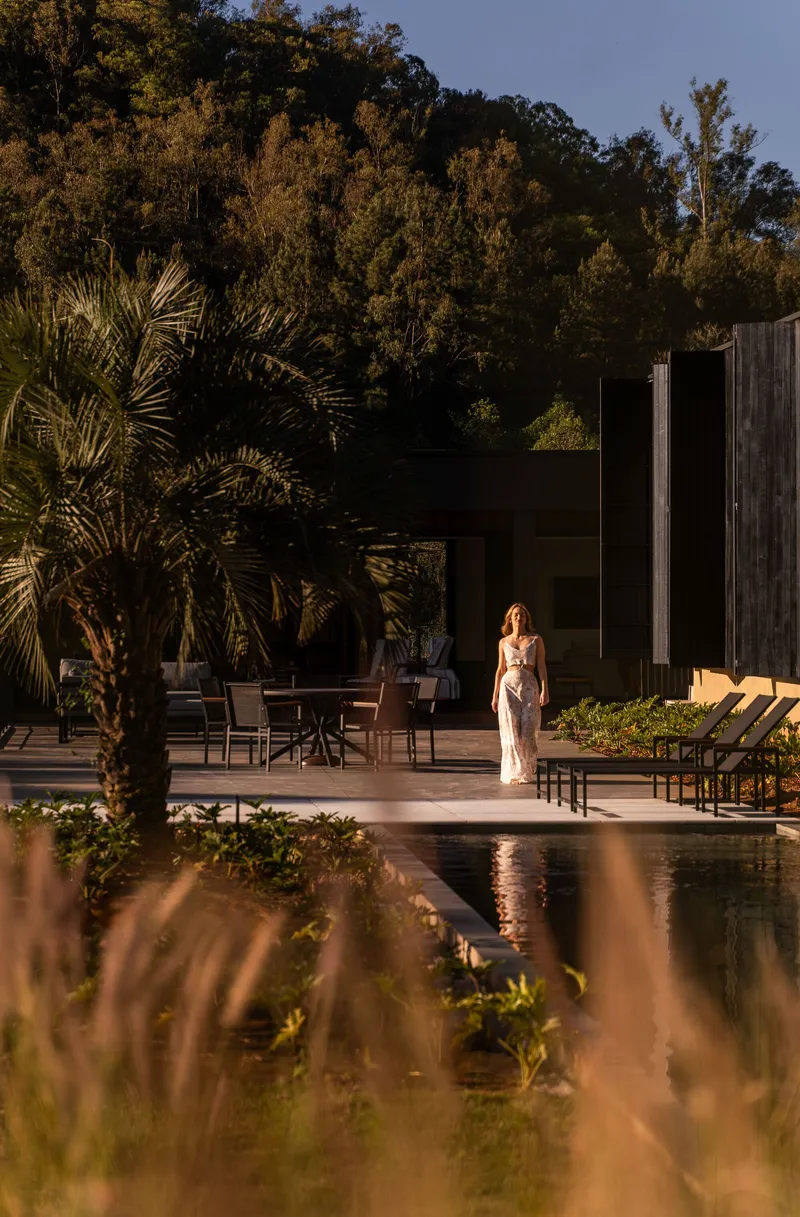 Poolside terrace at dusk with a figure standing near outdoor furniture and a palm tree