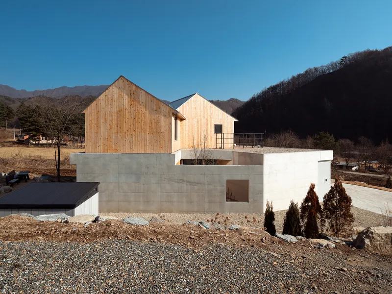 Timber-clad gabled volumes sitting atop a concrete base with gravel site and bare winter hills beyond
