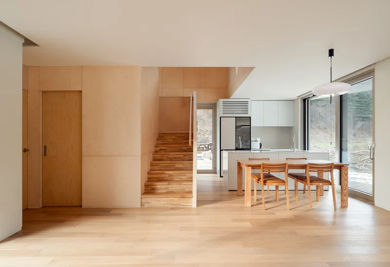 Open living and dining space with plywood stairs, white kitchen cabinetry, and timber table and chairs