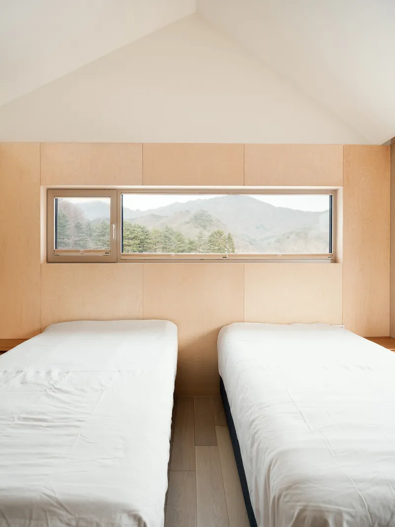 Bedroom with two single beds, plywood wall panels, and horizontal window opening to misty mountains