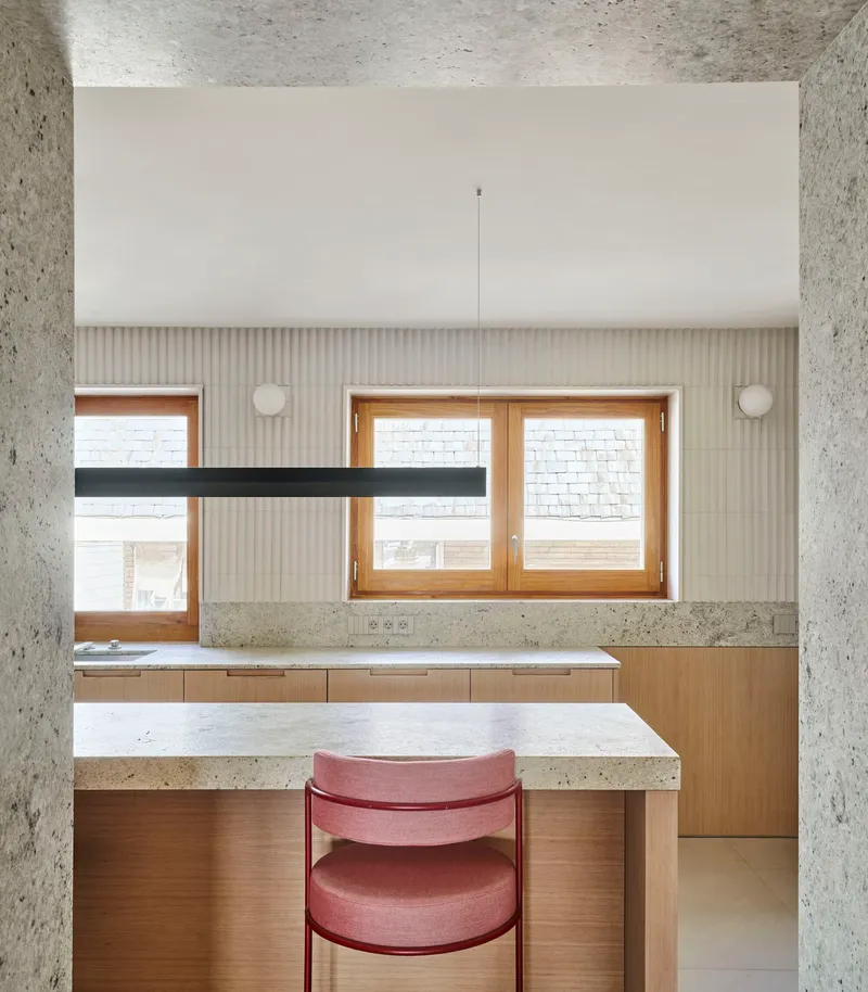 Kitchen workspace with terrazzo stone countertop, fluted wall cladding, and timber windows framing brick buildings