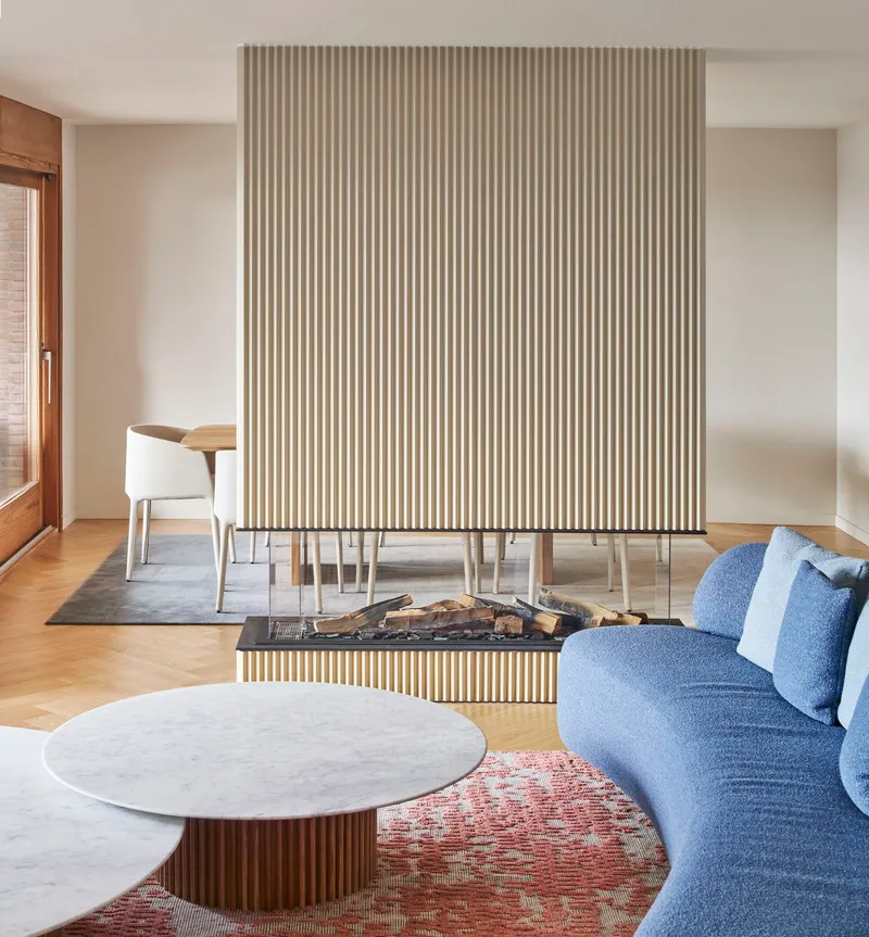 Fireplace with fluted surround holding stacked firewood, flanked by dining table and curved sofa