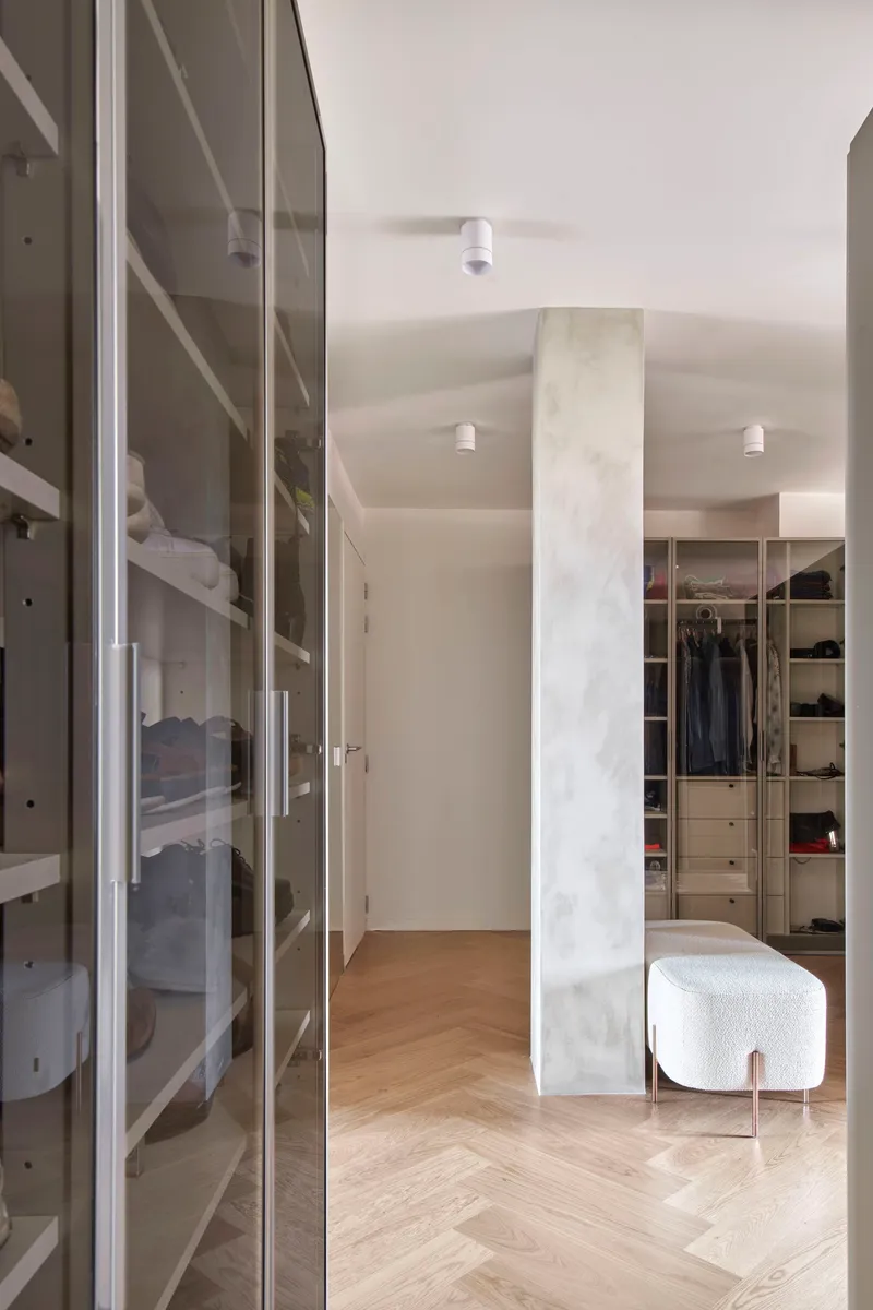 Walk-in closet with glass-fronted storage and white upholstered ottoman on herringbone oak flooring