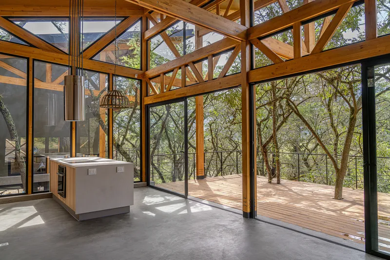 Kitchen with concrete island opening through floor-to-ceiling glazing onto a timber deck surrounded by trees