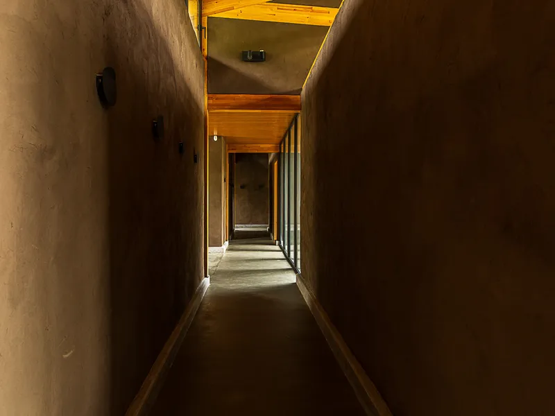 Narrow corridor with rammed earth walls and timber ceiling illuminated by recessed lights and end window