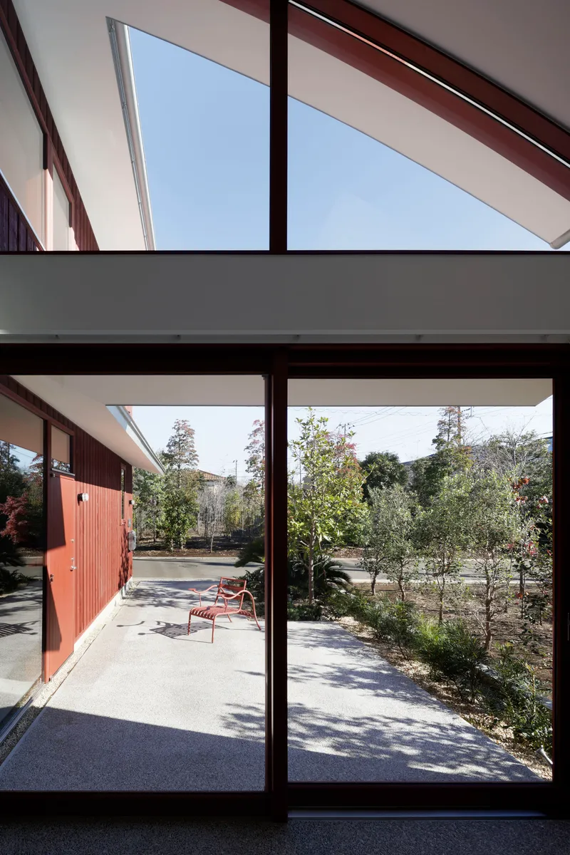 View through glazed doors to courtyard garden with planted trees and red bench under midday sun