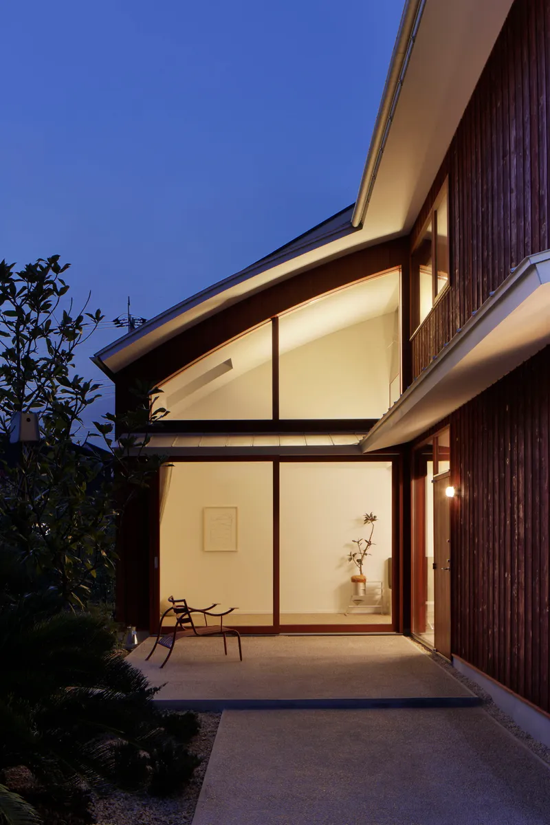Two-story glazed facade with timber cladding illuminated from within at dusk beside planted trees