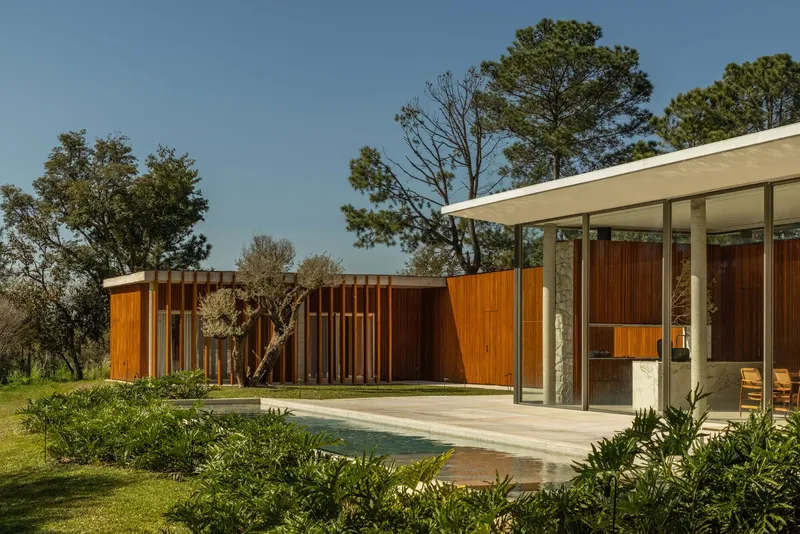 Covered terrace with stone columns and thin roof plane beside vertical timber screen walls in a grassy clearing