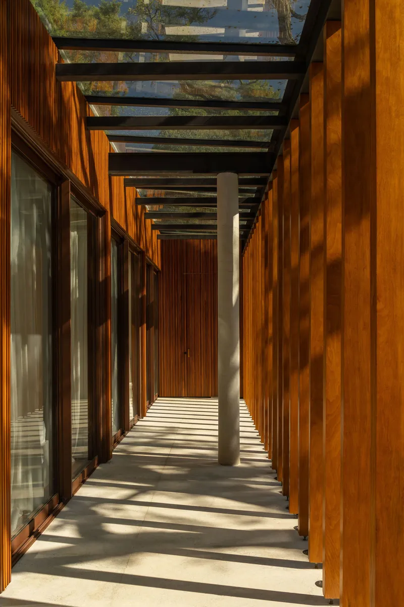 Covered walkway with timber slat walls casting striped shadows on pale stone floor beneath steel beams