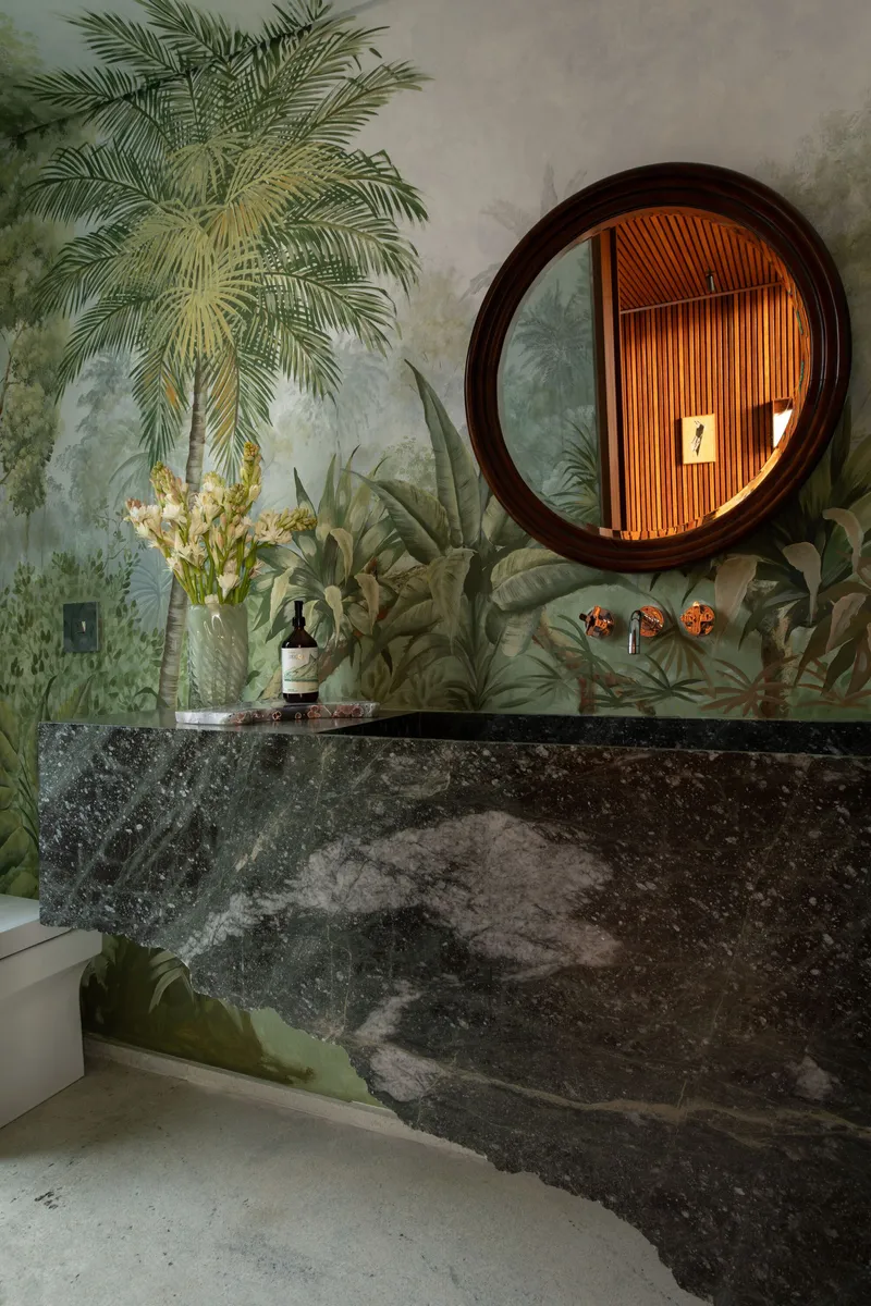 Bathroom vanity with dark stone countertop and circular mirror reflecting timber slats against botanical wallpaper
