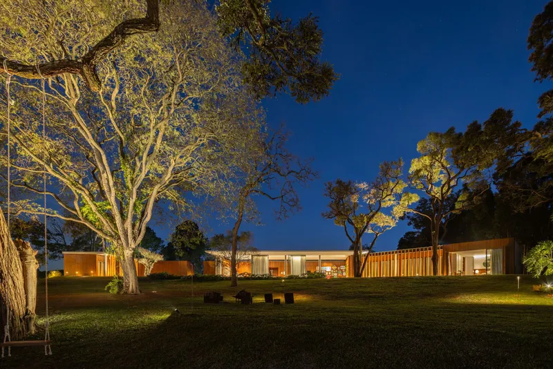 Low horizontal residence with timber screens and glazing nestled among uplighted trees at twilight