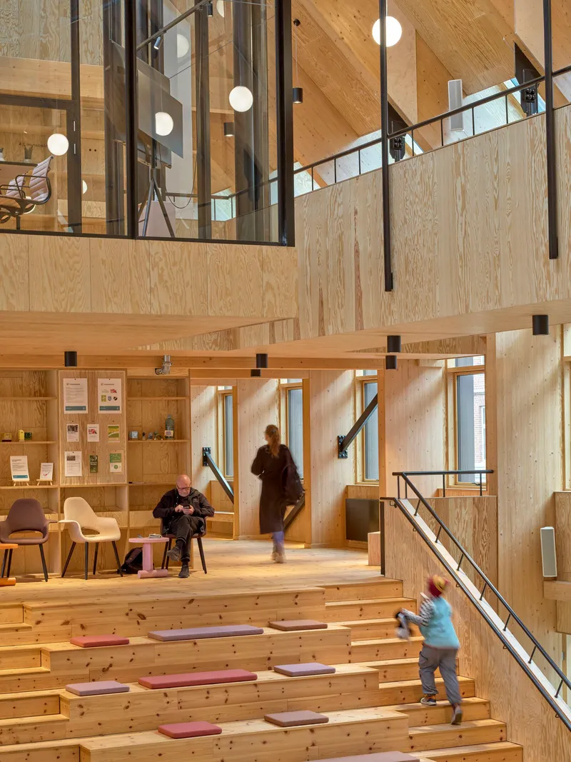 Tiered seating area with plywood benches and visitors moving through the double-height plywood-lined space