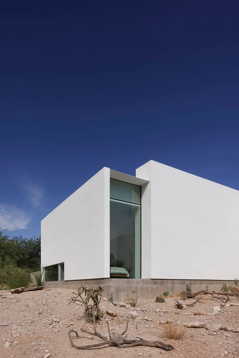 White stucco volumes framing a tall glazed slot window above desert terrain with scattered cacti