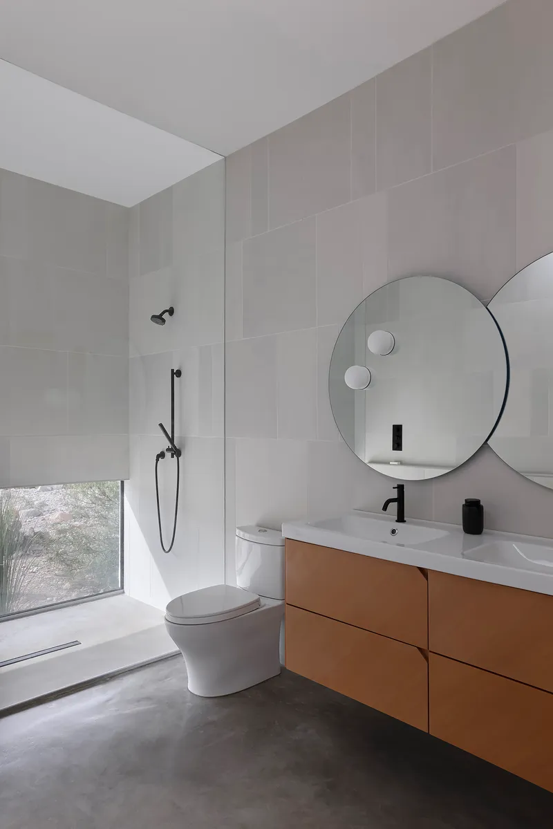 Bathroom with circular mirror above timber vanity and low desert window with roller shade