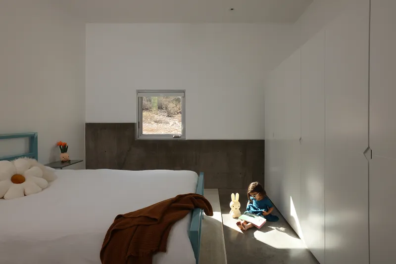 Child seated in sunlit corner beside bed with metal wainscoting and square desert window