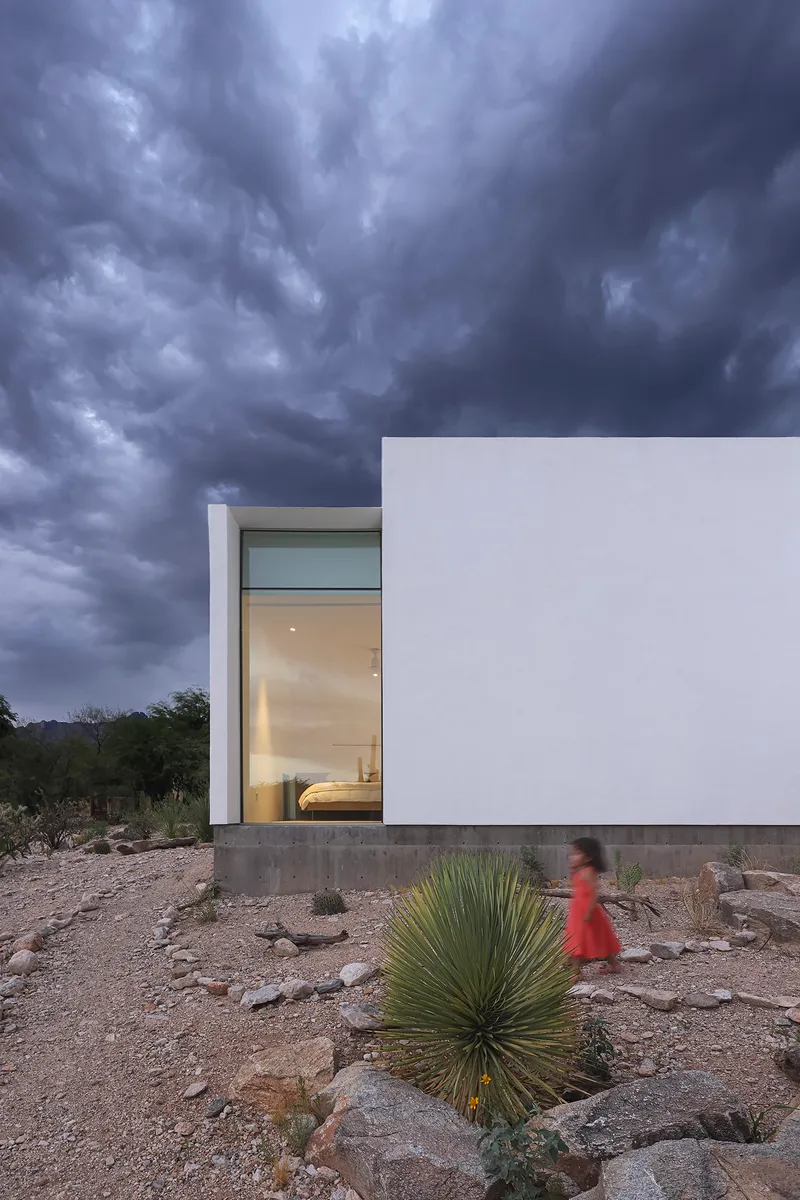 White stucco facade with recessed window opening and concrete base in desert landscape under storm clouds