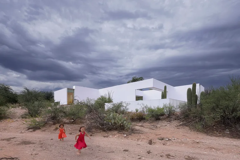 White geometric volumes with flat roofs and cutouts amid desert scrub with two children playing