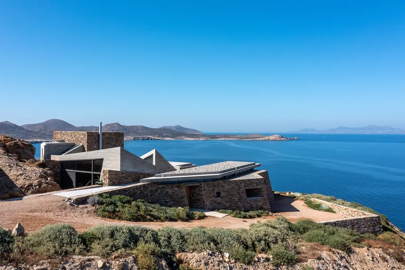 Coastal complex with stone volumes and white cantilevered roof overlooking a blue bay and distant mountains